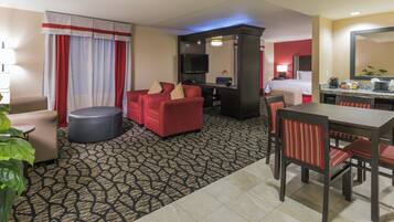 A hotel room with a living area featuring a red sofa, a round ottoman, a flat-screen TV, a dining table with chairs, and a kitchen area with a microwave and coffee maker.