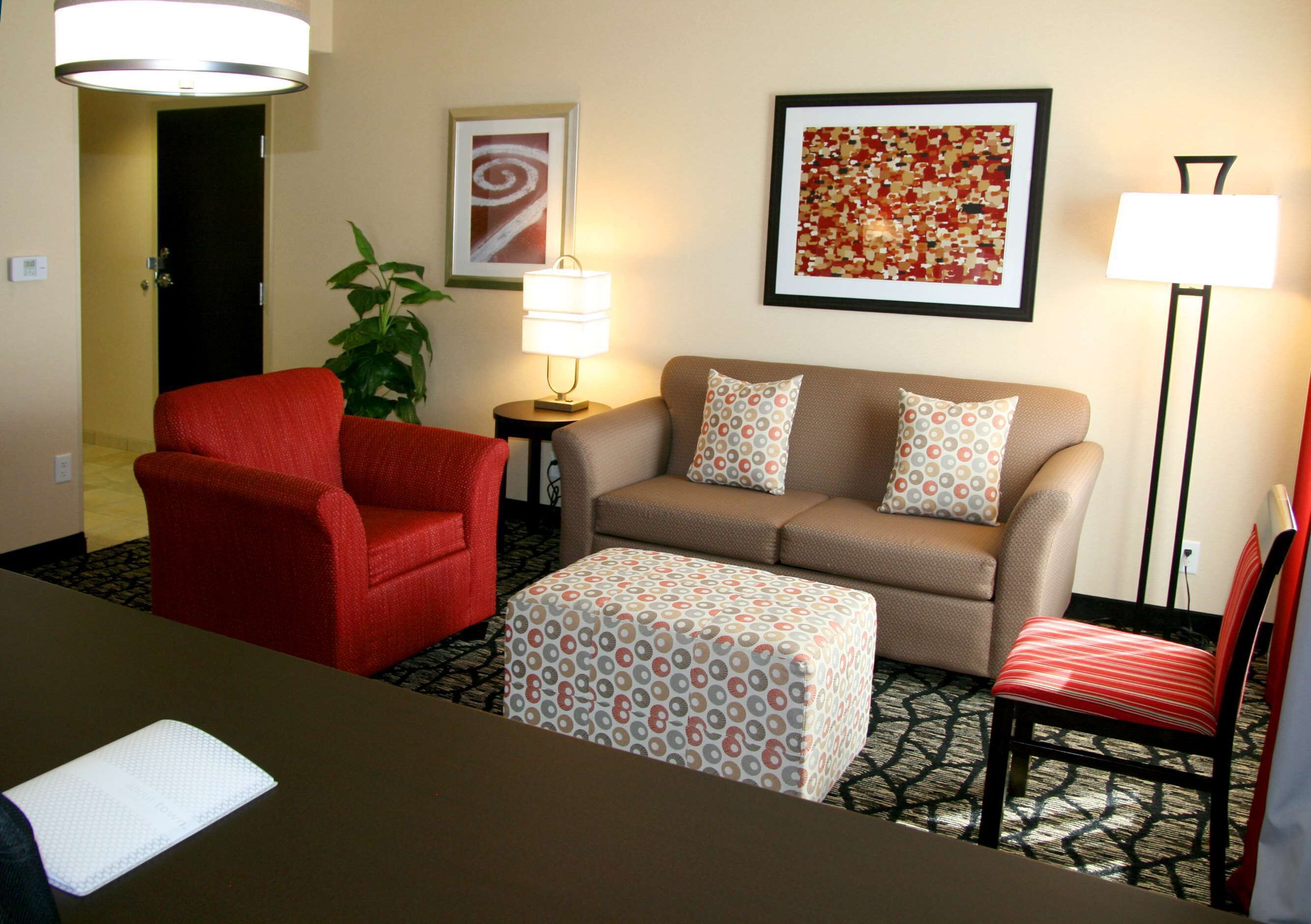 Habitación de hotel con un sillón rojo, un sofá color crema, un reposapiés estampado, una mesita con lámpara y un cuadro abstracto enmarcado en la pared.
