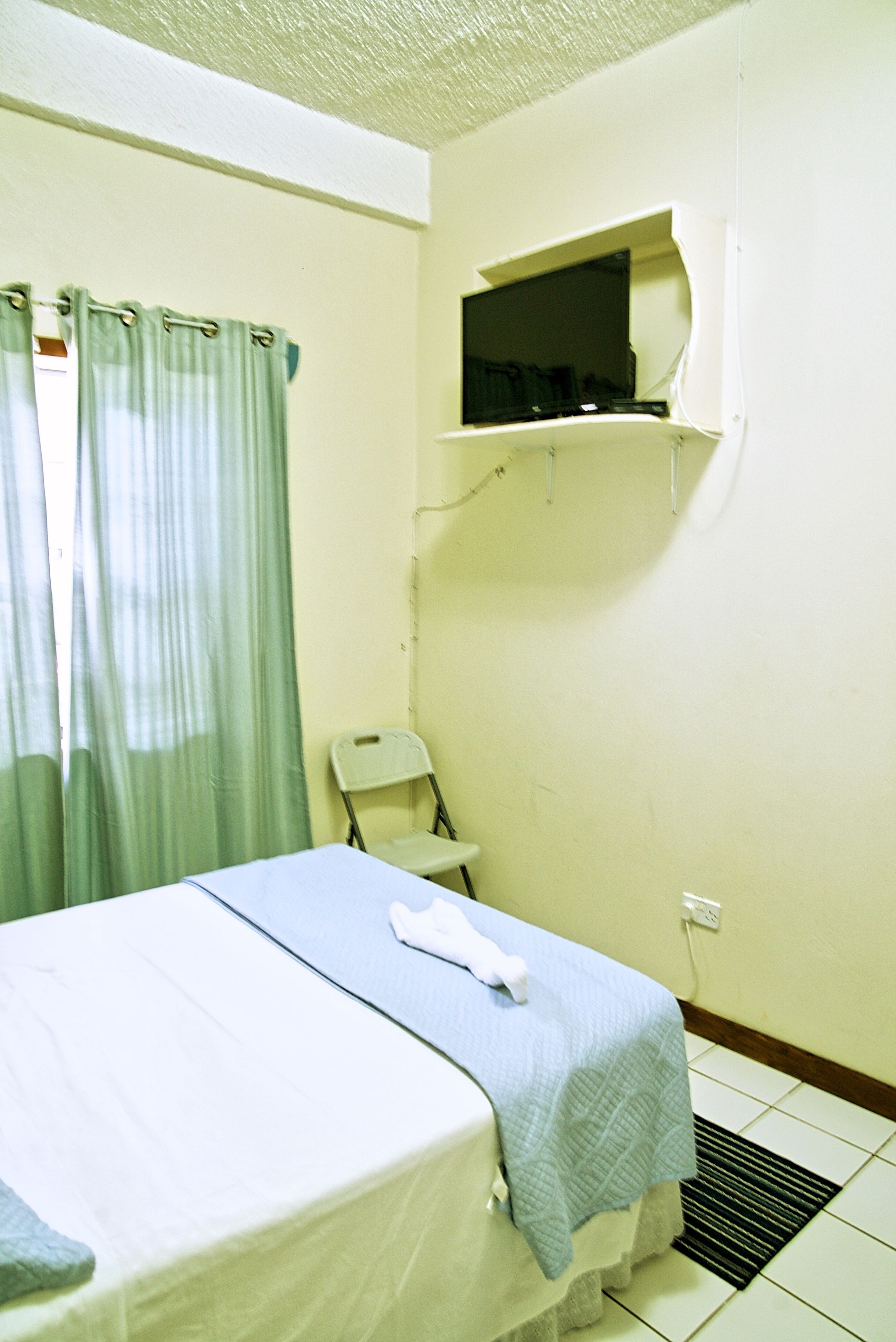 Una habitación de hotel pequeña con una cama, una silla, una ventana con cortinas y un estante sobre la cama.