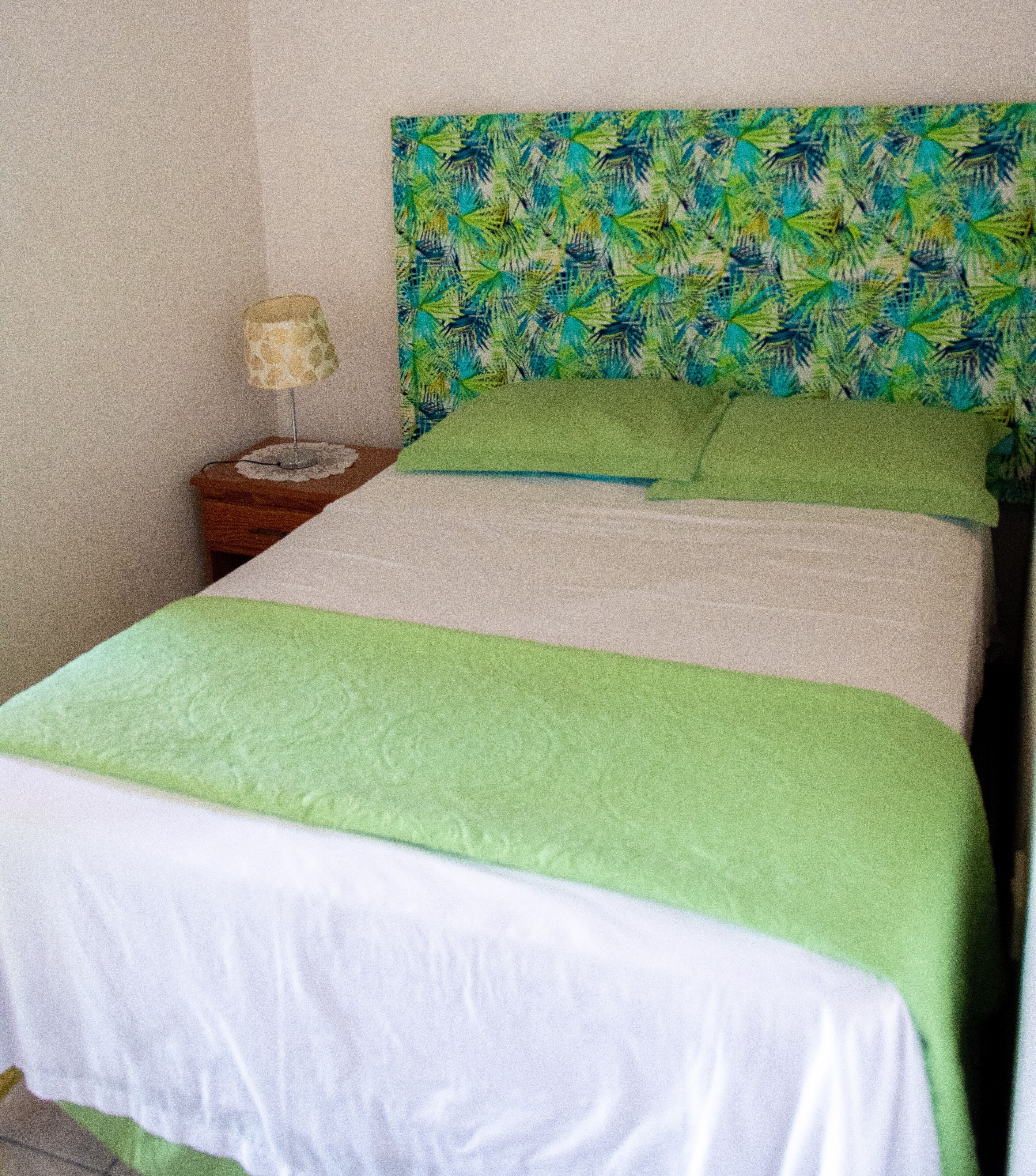 Una cama con cubrecama y almohadas verdes, una mesita de noche con lámpara y una pared con estampado de hojas tropicales.
