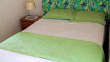 Una cama con cubrecama y almohadas verdes, una mesita de noche con lámpara y una pared con estampado de hojas tropicales.