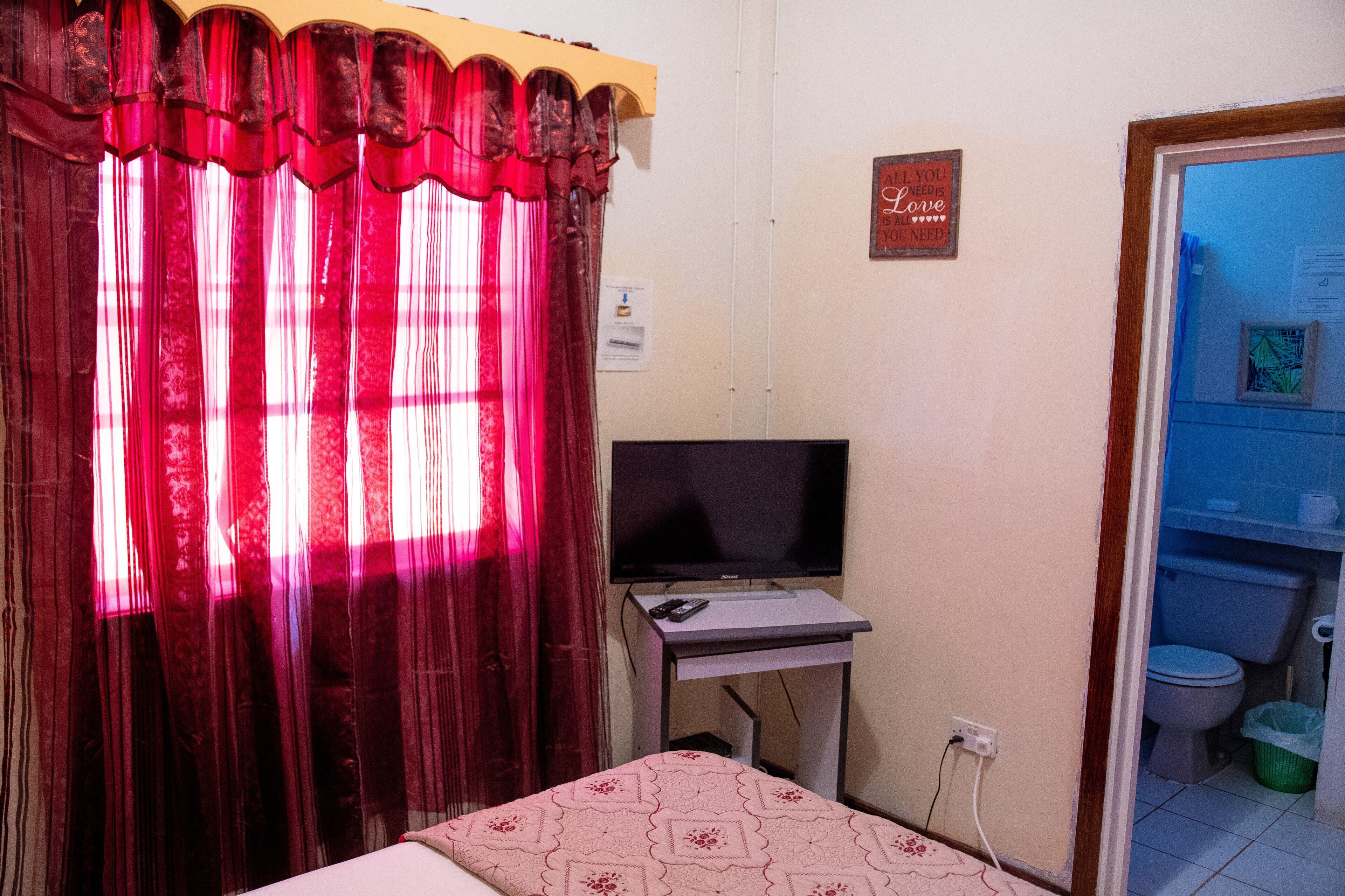 A small room with a bed, a television on a stand, a curtain, a wall-mounted sign, and a door leading to a bathroom.