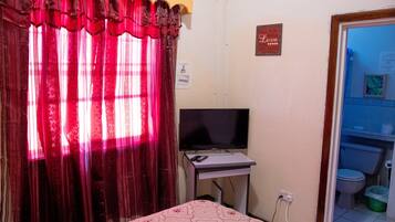 A small room with a bed, a television on a stand, a curtain, a wall-mounted sign, and a door leading to a bathroom.