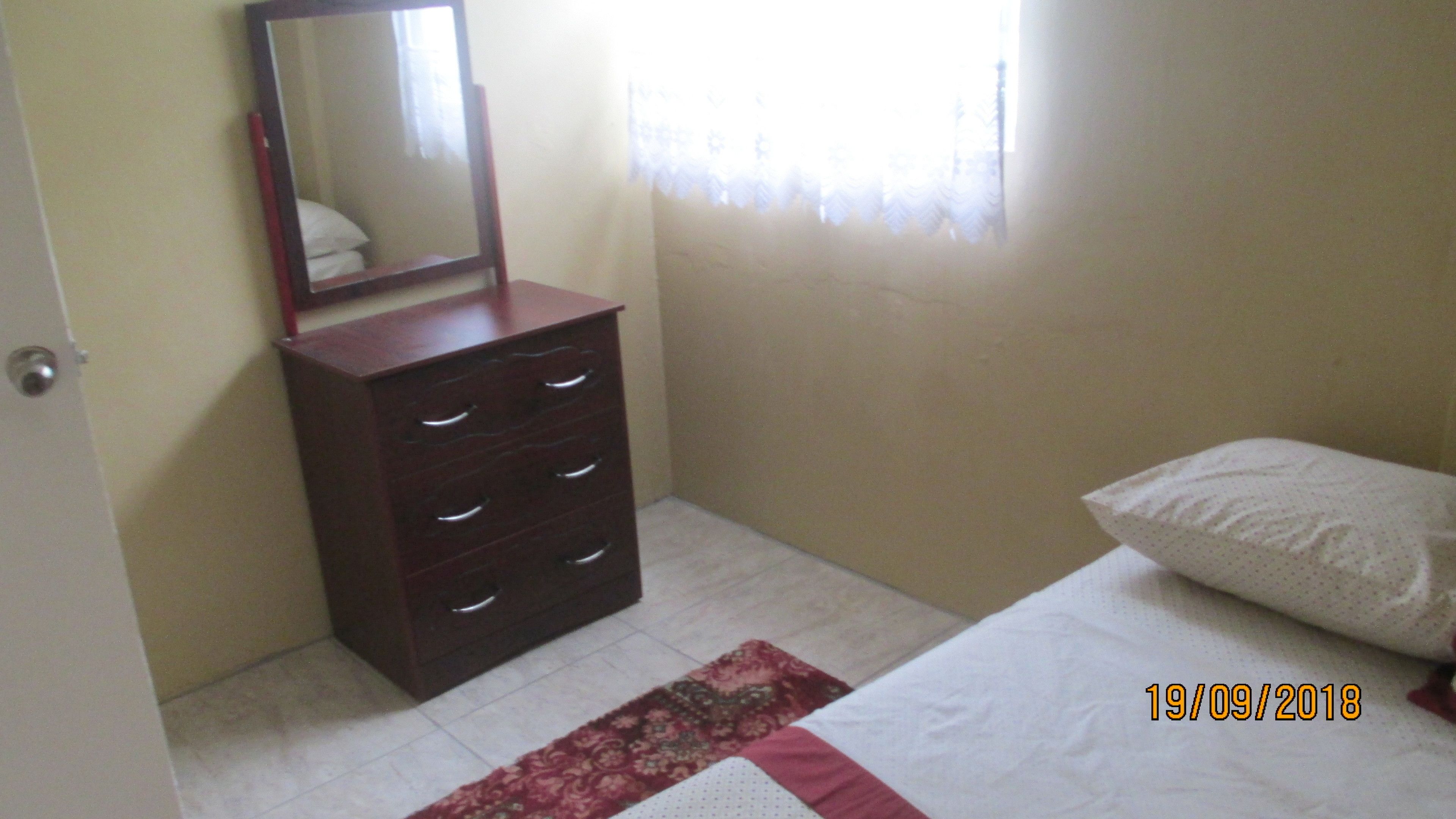 A bedroom with a bed, a dresser with drawers, a mirror, a window with curtains, and a door.