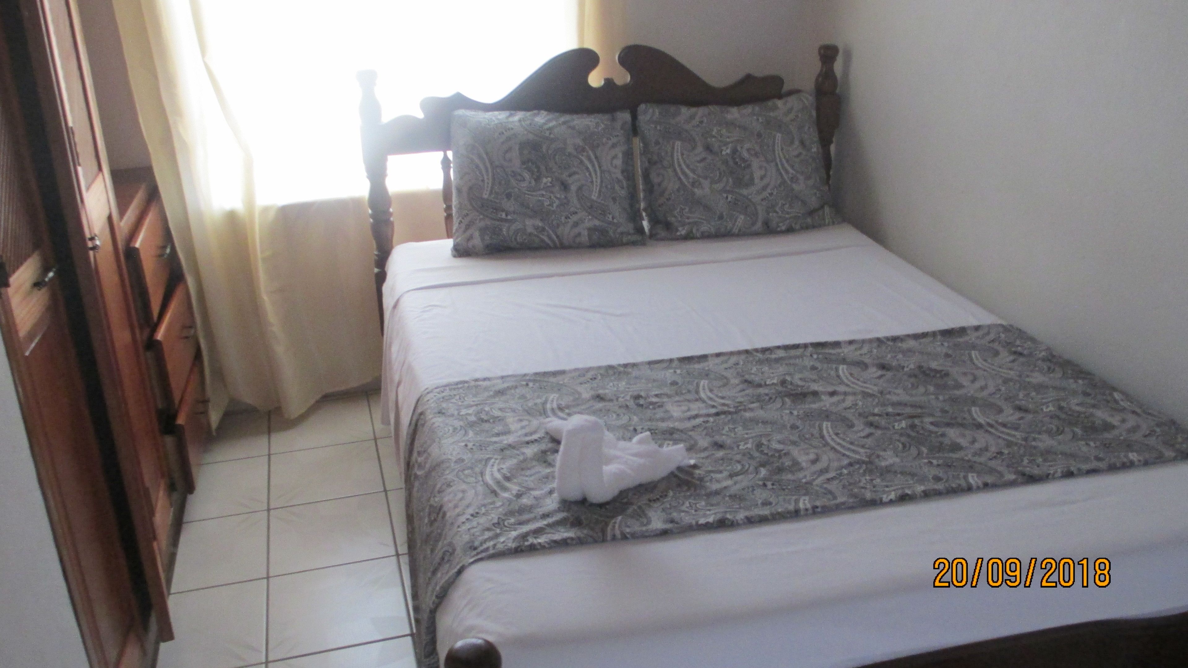 A neatly made bed with a patterned bedspread and pillows, a wooden headboard, and a wooden dresser to the left.