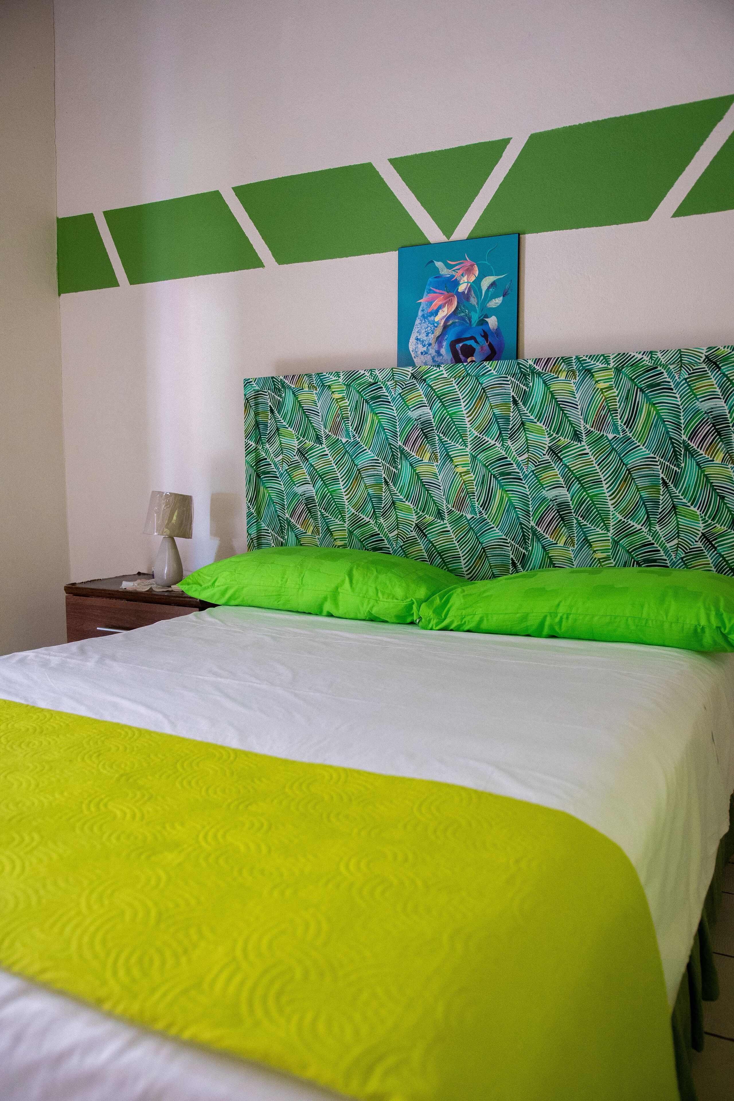 Uma cama com colcha verde e branca, um travesseiro verde e um quadro de um peixe na parede acima da cama.