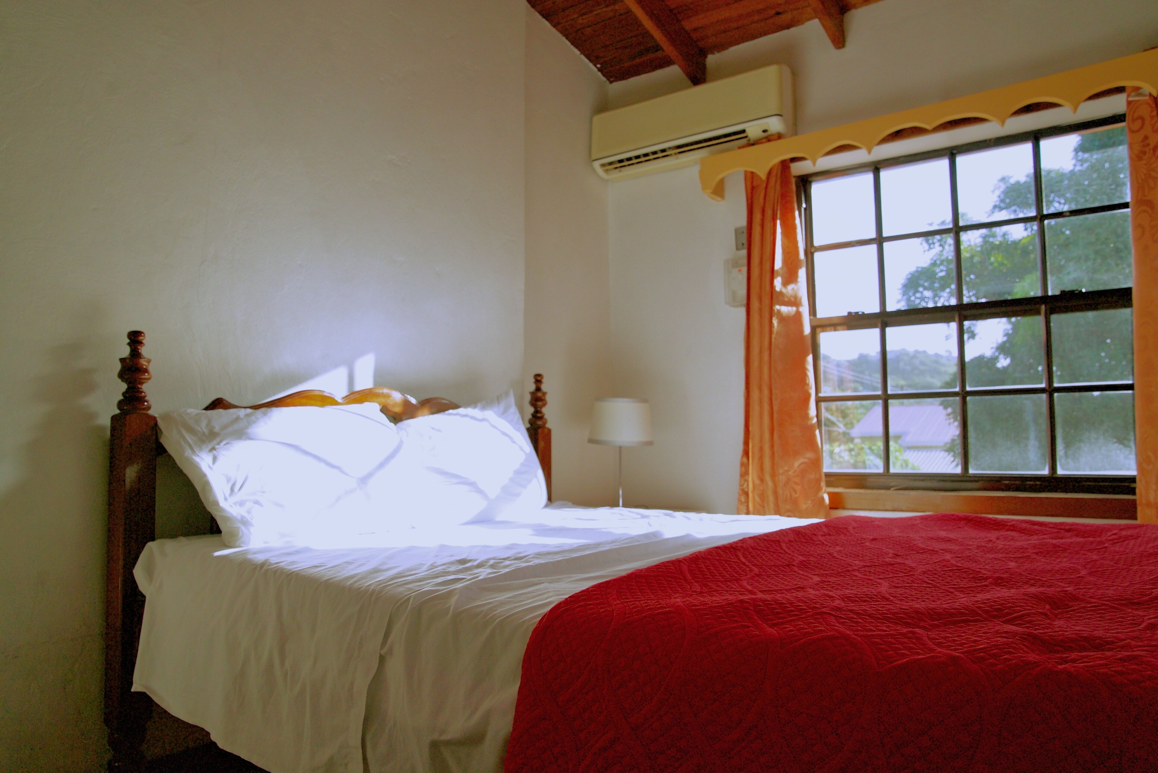 A bed with a wooden headboard, white bedding, and a red blanket. There is a window with orange curtains and a view of trees outside.