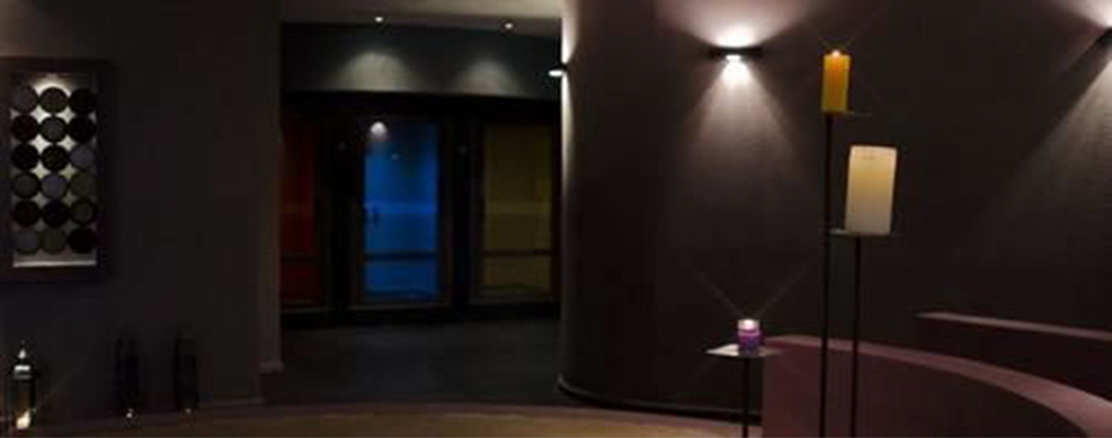 A dimly lit hallway with a circular wall design, a door leading to a blue-lit room, and a small table with a candle.