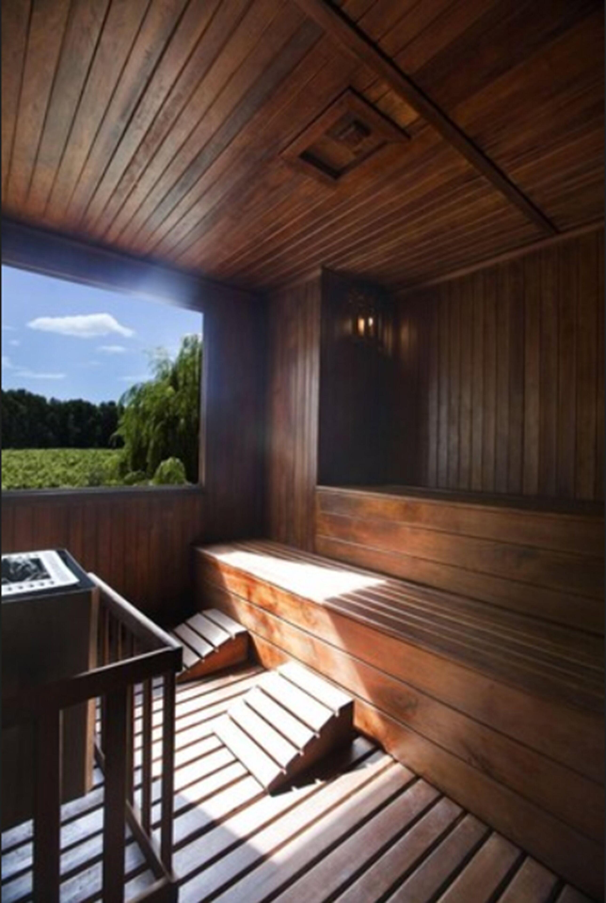 A sauna with wooden walls and a bench, a window with a view of greenery, and a table with a chair.