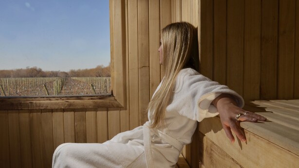 A person in a white bathrobe sitting by a wooden window with a view of a wooden deck. - Fuente Mayor Hotel And Resort (Tunuyan)