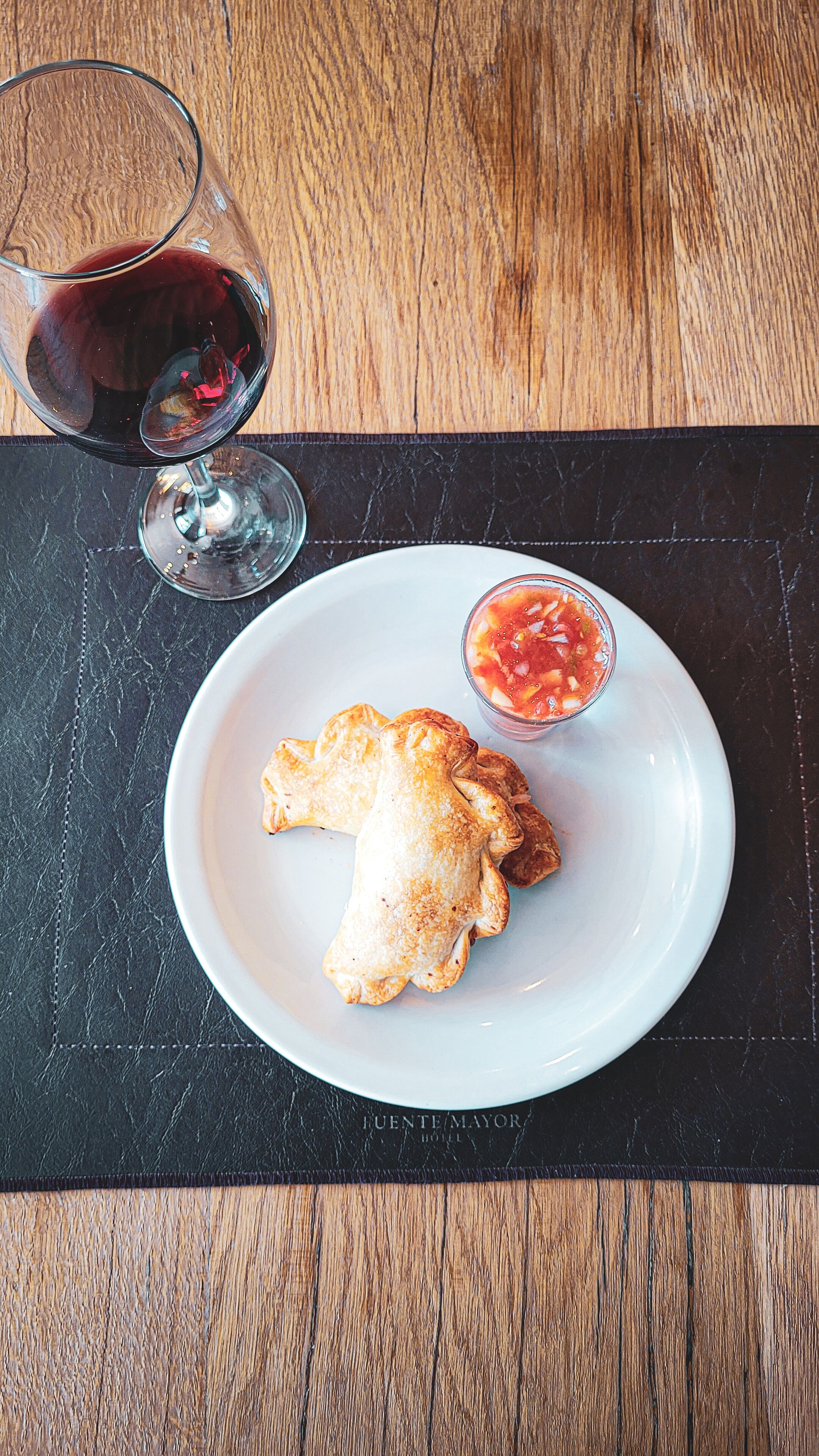 A plate with two pastries and a small bowl of salsa, accompanied by a glass of red wine on a wooden table.