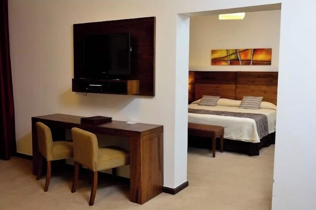 A hotel room with a bed, a desk with two chairs, a television mounted on the wall, and a painting above the bed.