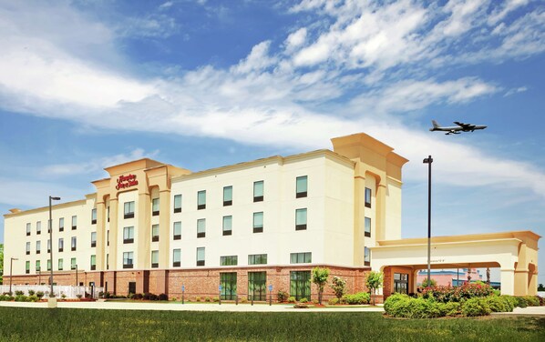 Ein Hampton Inn Hotel mit Backsteinfassade, zahlreichen Fenstern und blauem Himmel mit einem Flugzeug im Hintergrund.