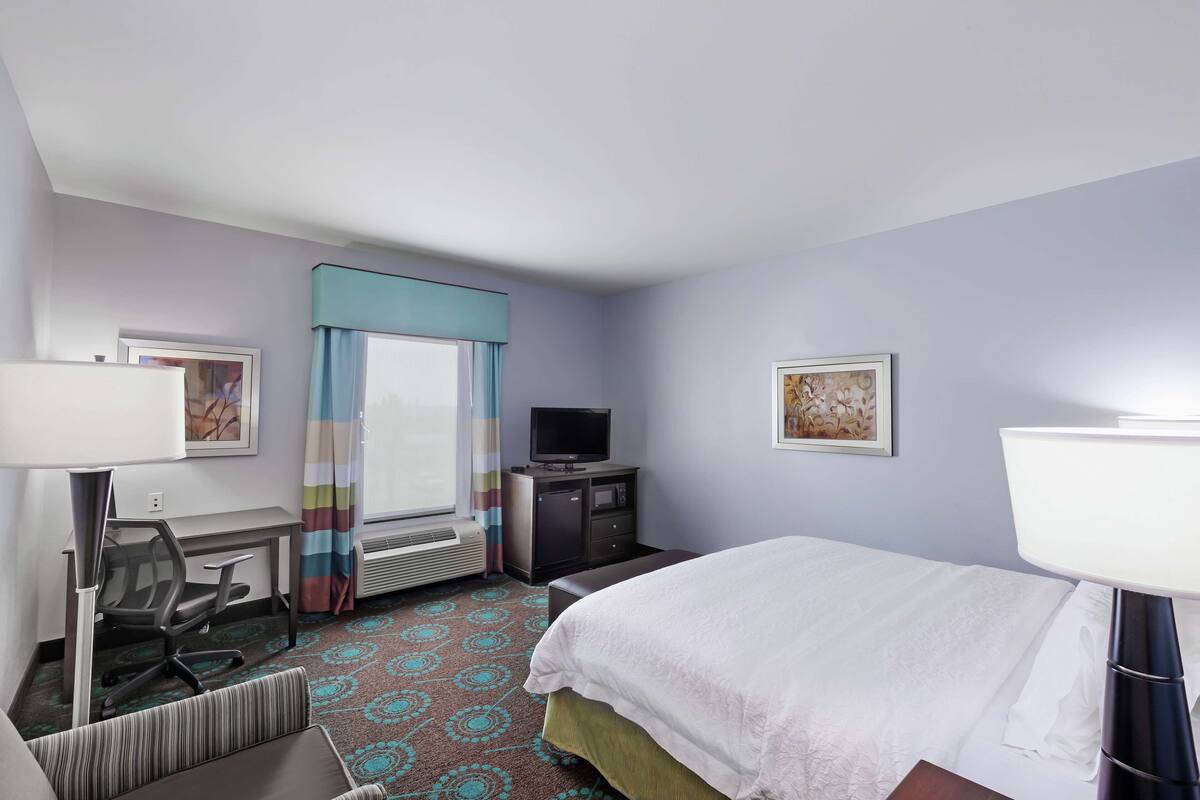 Une chambre d’hôtel comprenant un lit, un bureau avec une chaise, une télévision et une fenêtre avec des rideaux.