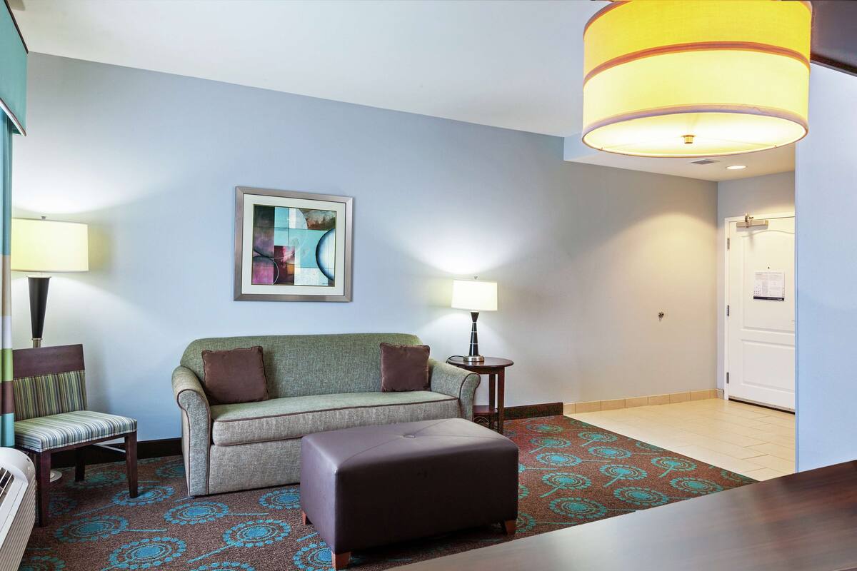 Une chambre d’hôtel avec un canapé, un fauteuil, une petite table, une lampe, un tableau au mur et une porte.