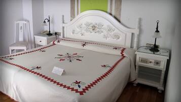 Een net opgemaakt bed met een bloemenbedekking en een rode, golvende rand, twee nachtkastjes met lampen, een stoel en een raam met uitzicht op groen.