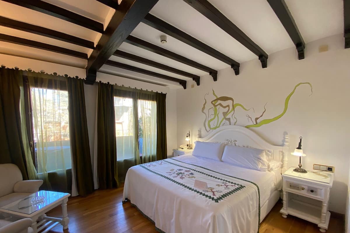 Ein Hotelzimmer mit einem großen Bett, einem Schreibtisch und einem Stuhl. Es gibt ein Fenster mit Vorhängen, eine Wand mit einem dekorativen Gemälde und einen Nachttisch mit einer Lampe.