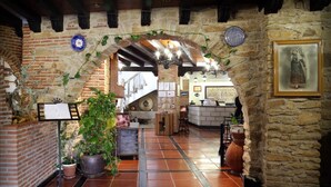 A rustic interior with exposed brick walls, a tiled floor, and a wooden ceiling. There is a brick fireplace, a dining area with a table and chairs, and a counter with a plant. - Hotel Doña Teresa (La Alberca)