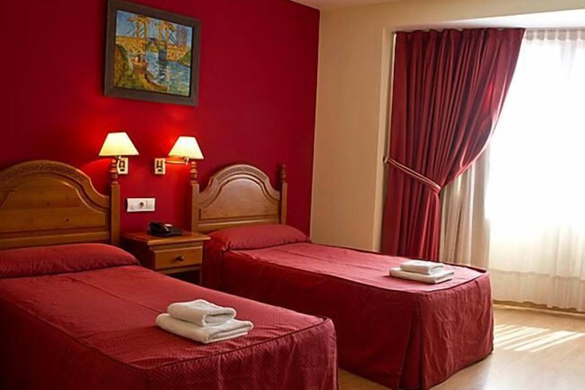 A hotel room with two single beds, red walls, a painting, and a window with red curtains.