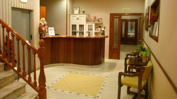 A hotel reception area with a wooden reception desk, a waiting area with a chair, a staircase, and a sign indicating a restaurant. - Hotel Clemente (Barbastro)