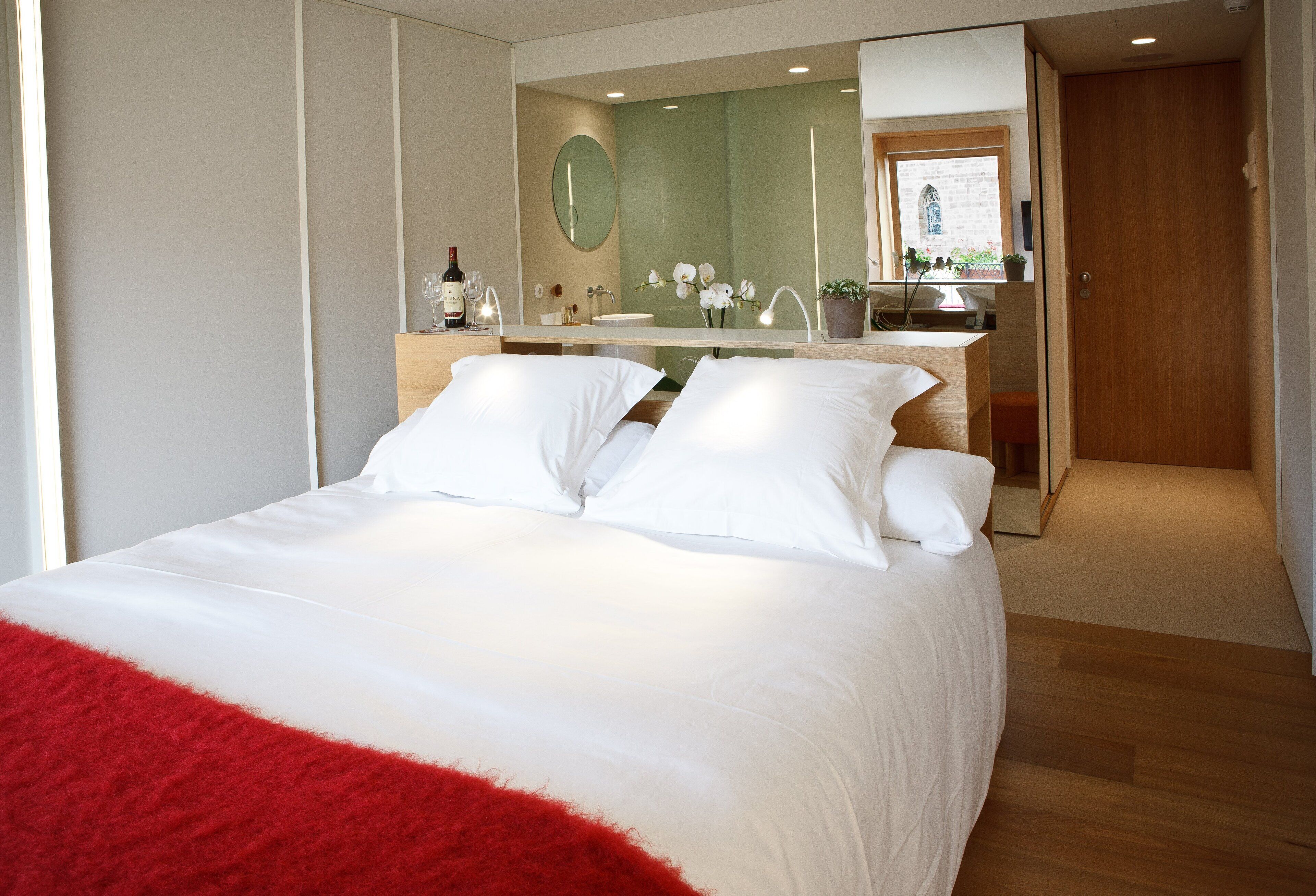Une chambre d’hôtel moderne avec un grand lit, une commode en bois, une coiffeuse avec un miroir et une petite fenêtre donnant sur un immeuble.