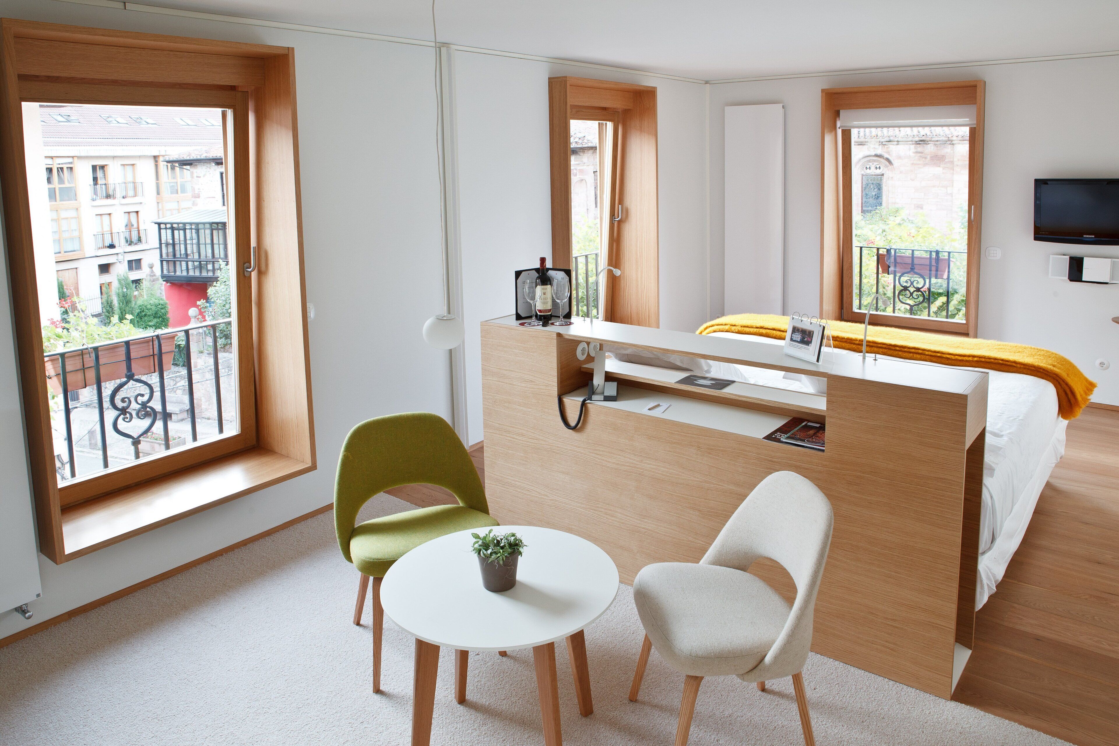 Habitación de hotel pequeña con cama, escritorio, dos sillas, una mesita con una planta y ventanas amplias con vistas a la ciudad.