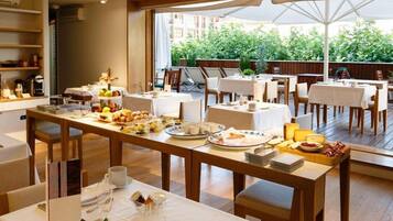 A dining area with tables set for breakfast, featuring a variety of food items and beverages, with a view of greenery outside.