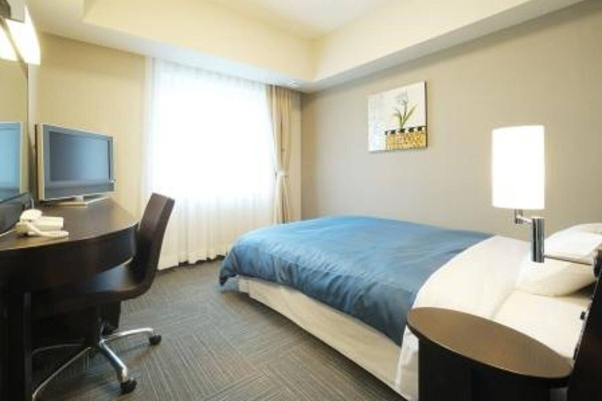 A hotel room with a bed, a desk with a computer, a chair, and a lamp.