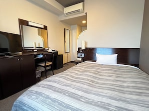 Ein Hotelzimmer mit Bett, Schreibtisch, Stuhl, Kommode, Spiegel, Nachttisch und Klimaanlage.