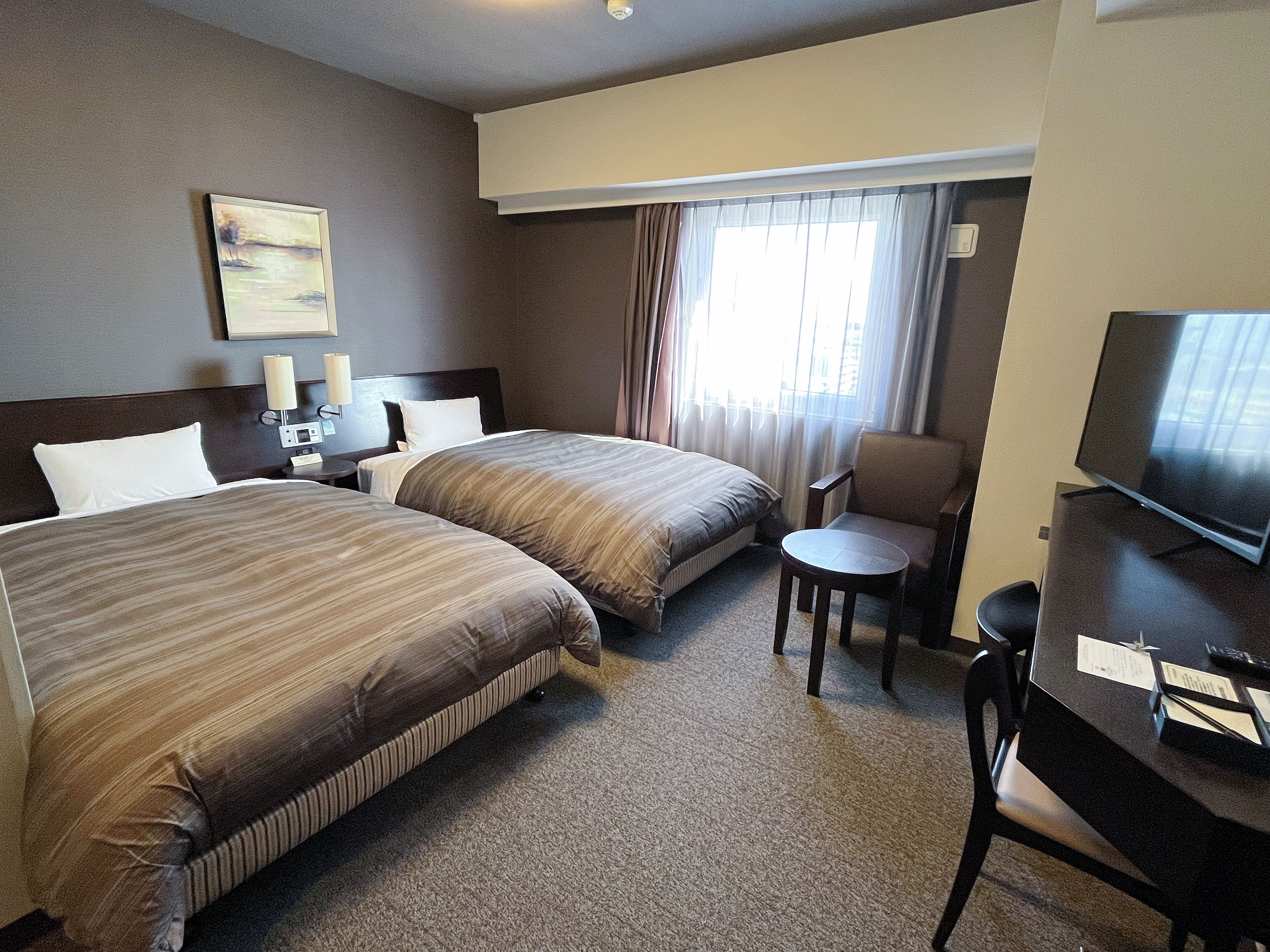 Ein Hotelzimmer mit zwei Betten, einem Schreibtisch mit Fernseher, einem Sessel, einem kleinen Tisch und einem Fenster mit Vorhängen.