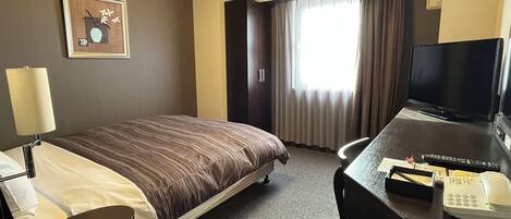 A hotel room with a bed, a desk with a television, a lamp, and a window with curtains.