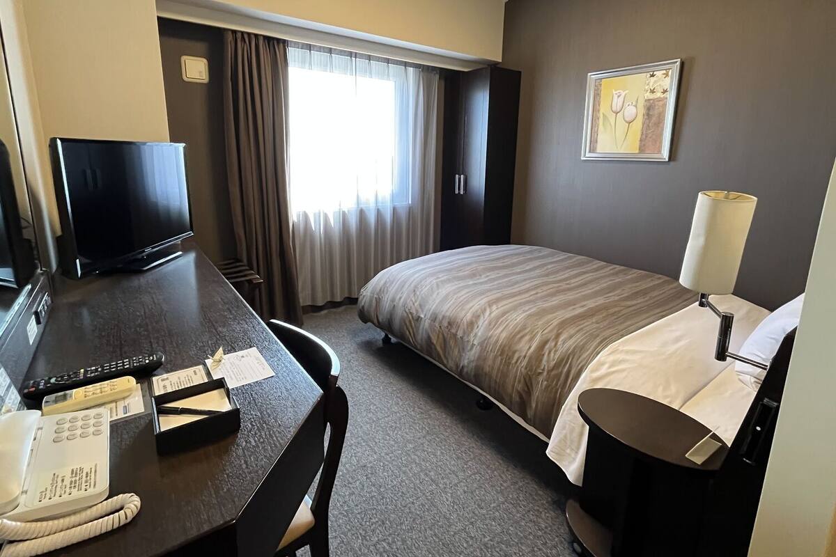 A hotel room with a bed, a desk with a telephone and remote controls, a television, and a lamp.