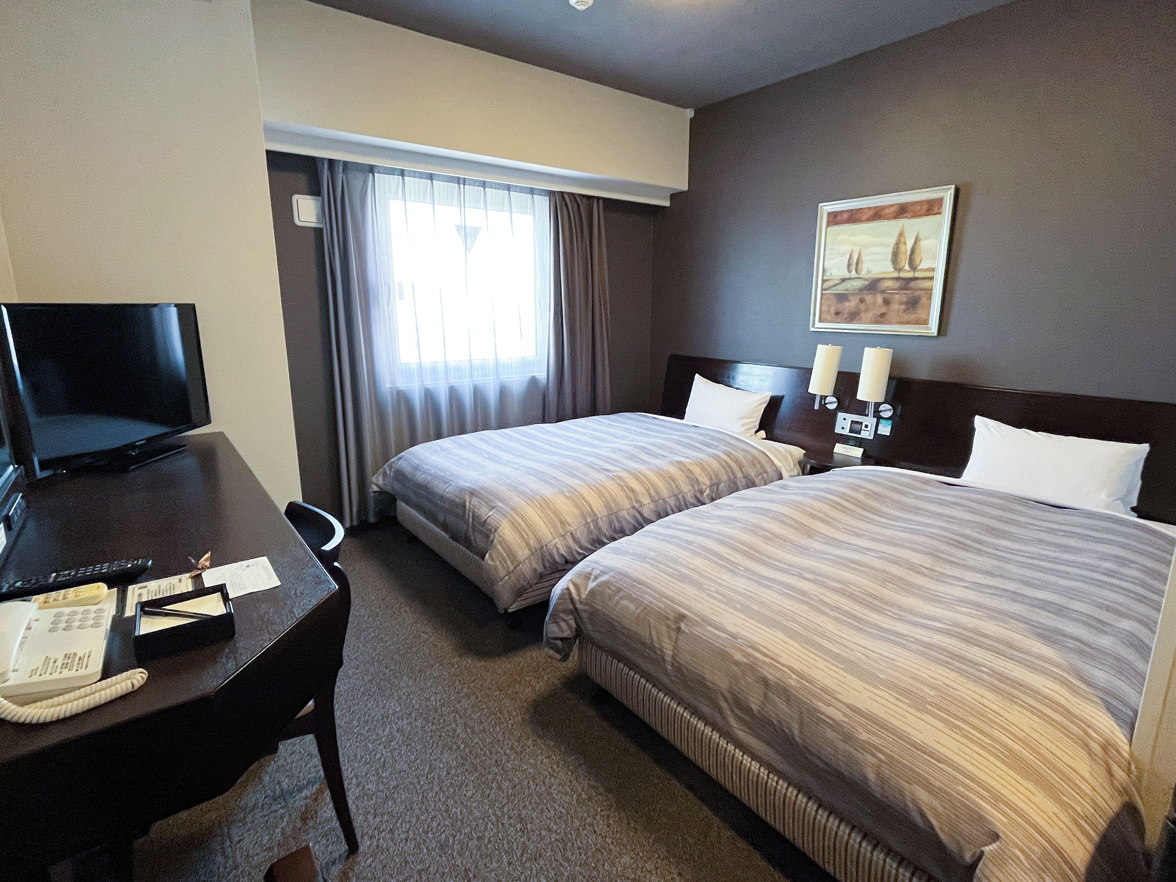 A hotel room with a bed, a desk with a TV, a telephone, and a painting on the wall.