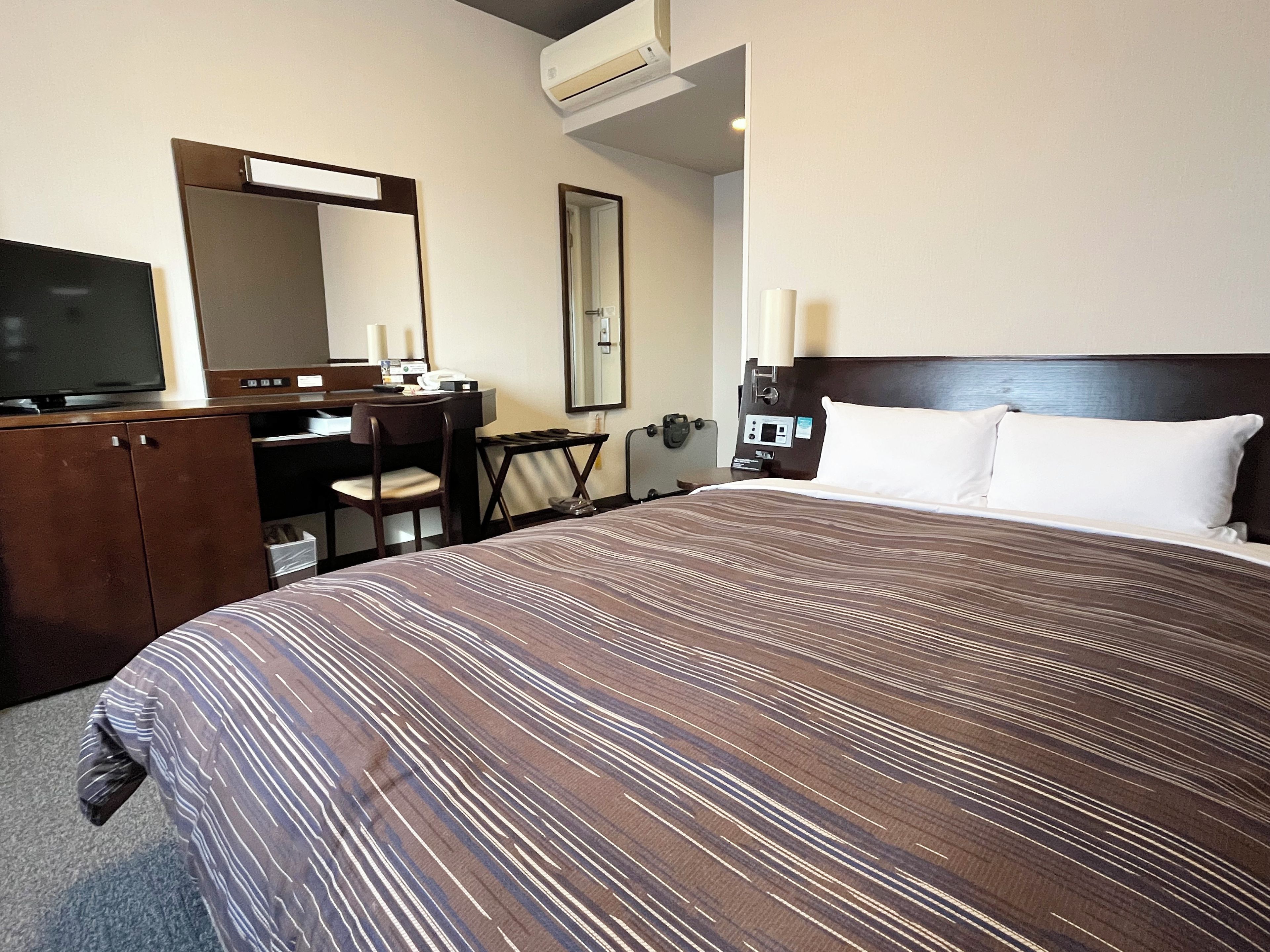 A hotel room with a large bed, a desk with a chair, a television, a mirror, and a bedside table with a lamp.