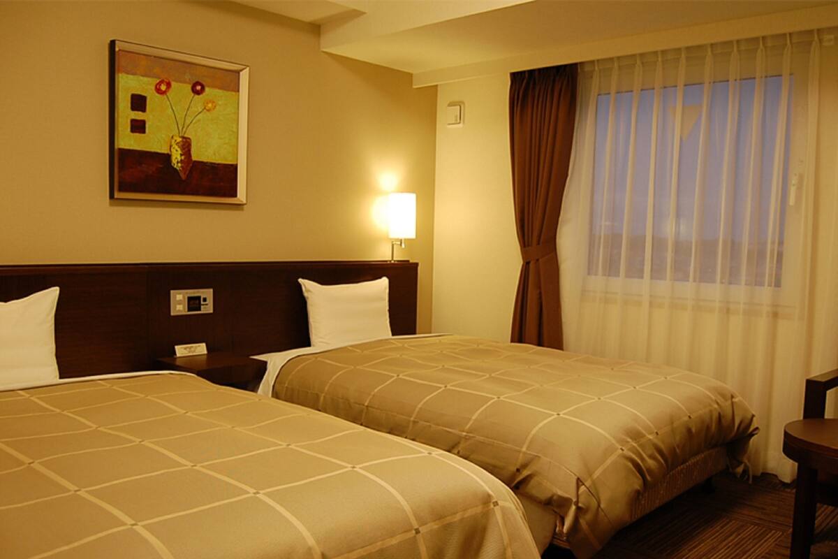 A hotel room with two beds, a painting on the wall, a nightstand, a lamp, and a window with curtains.
