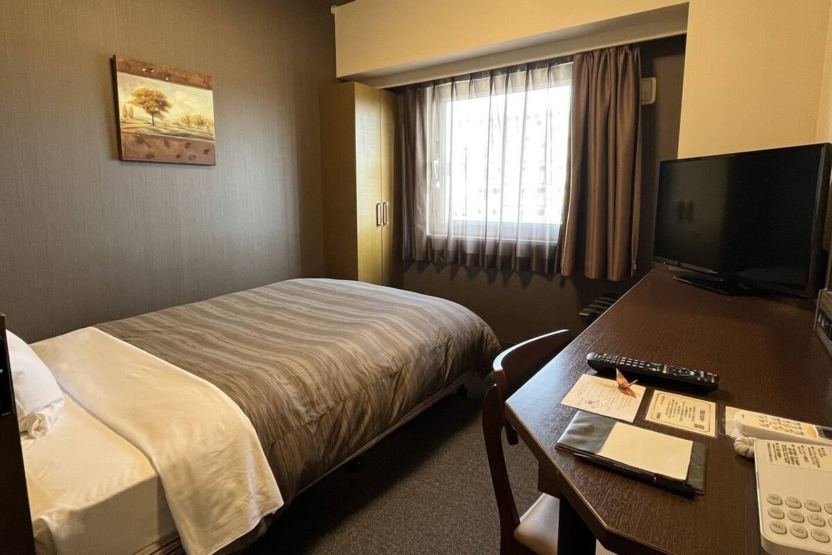 A hotel room with a bed, a desk with a TV, a remote, and some papers.