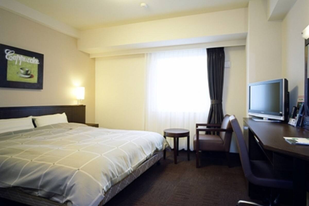 A hotel room with a bed, a desk with a chair, a television, and a window with curtains.