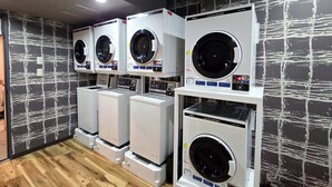 A row of four front-loading washing machines in a showroom with a wooden floor and a black and white abstract wall design. - Hotel Route-Inn Dai-Ni Nagano (Nagano)