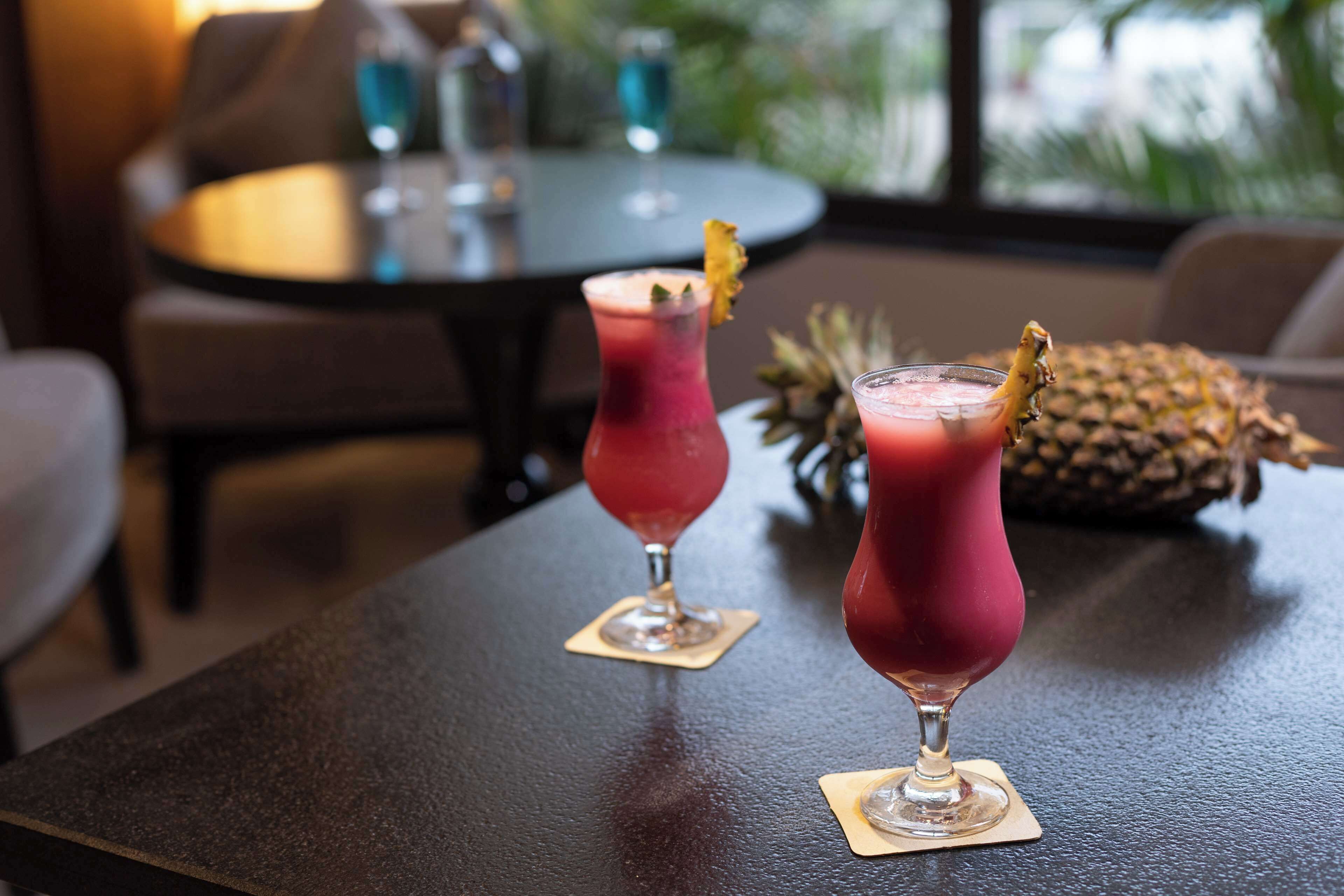 Dos bebidas sobre una mesa, con una piña y una botella al fondo.