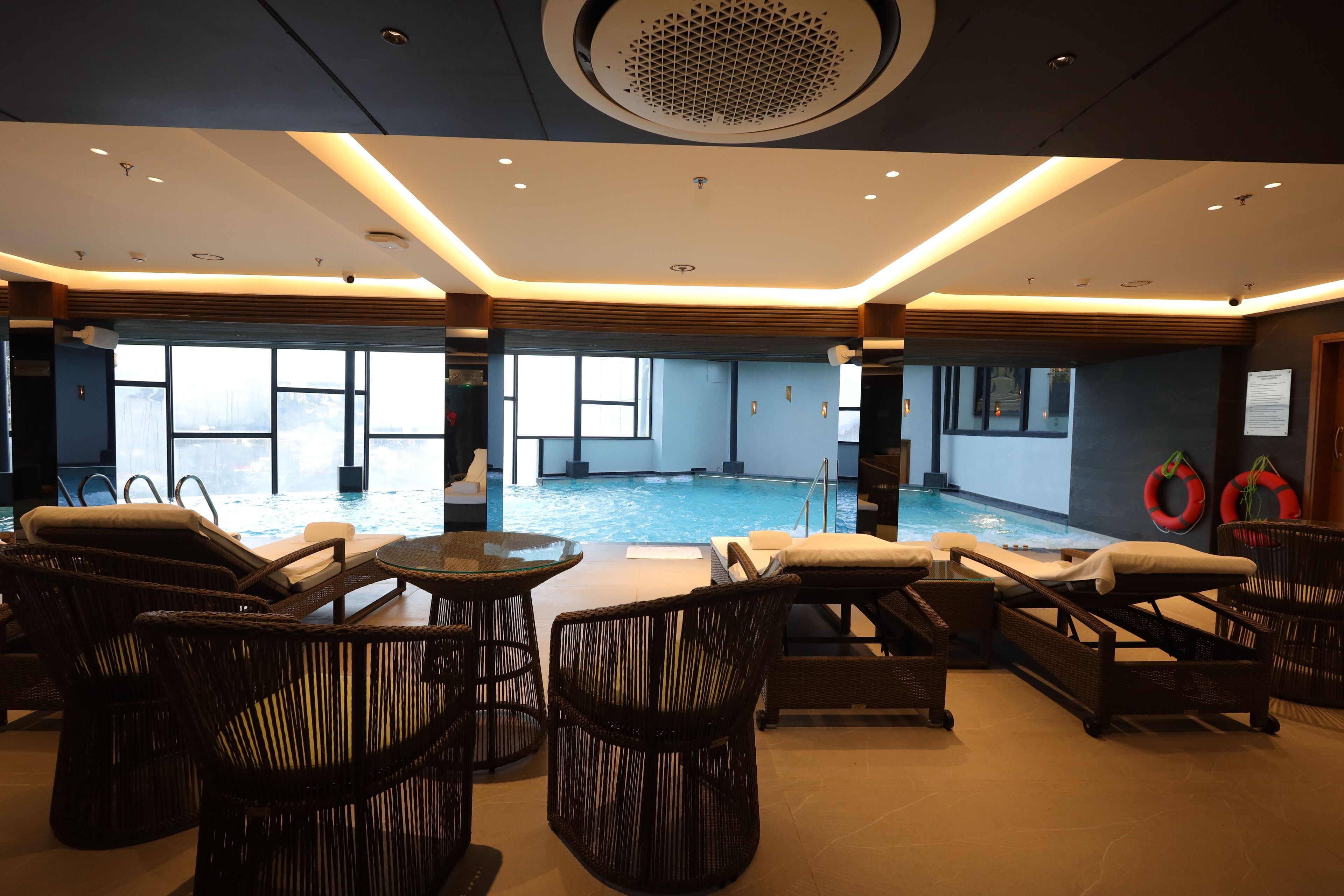 A modern indoor pool area with lounge chairs and tables, large windows, and a ceiling with recessed lighting.