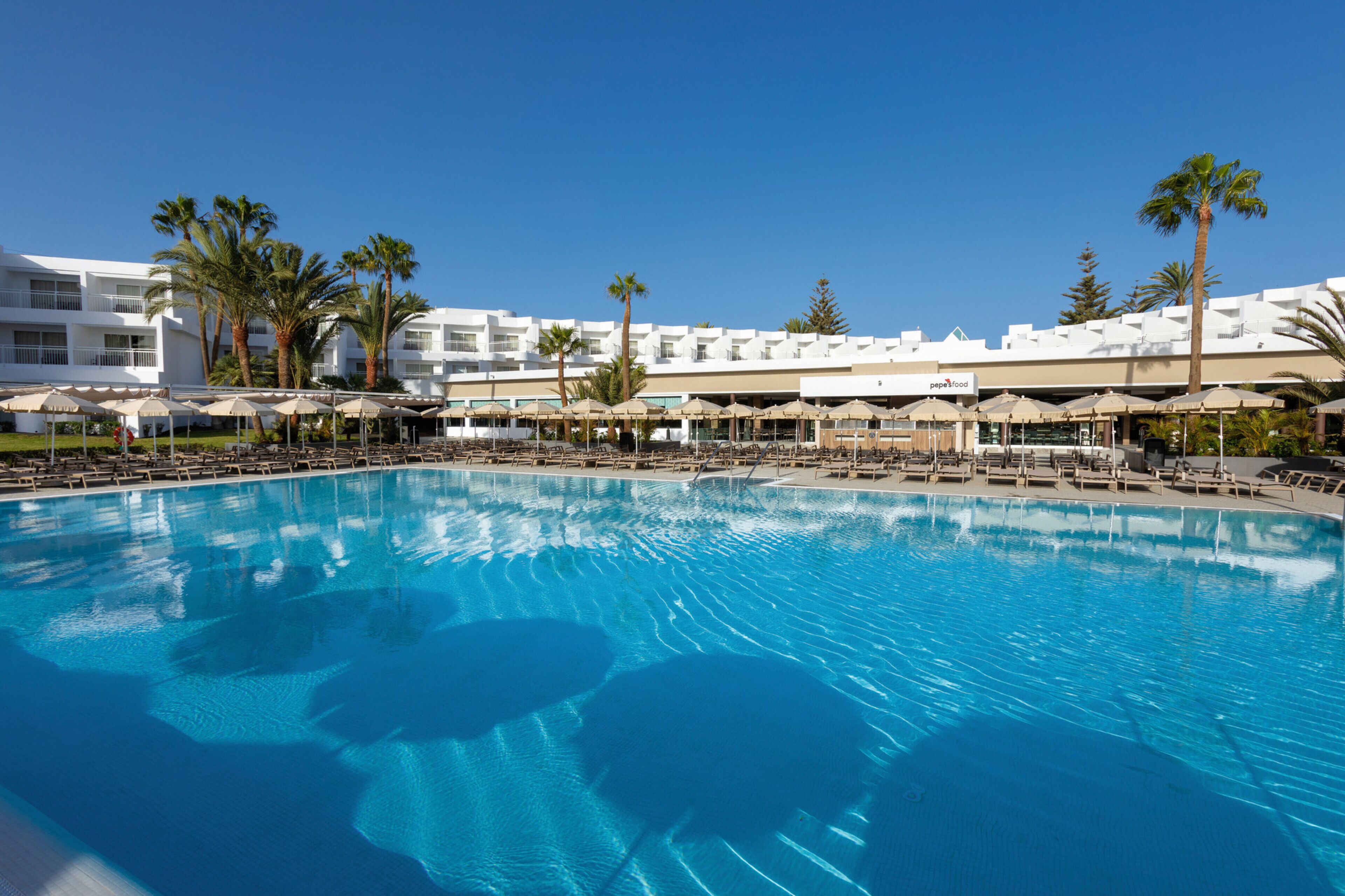 A large pool with lounge chairs and umbrellas, surrounded by palm trees and a hotel building.
