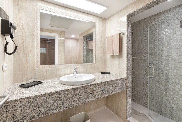 A modern bathroom with a granite countertop, a white sink, a large mirror, a hairdryer, and a glass shower enclosure.
