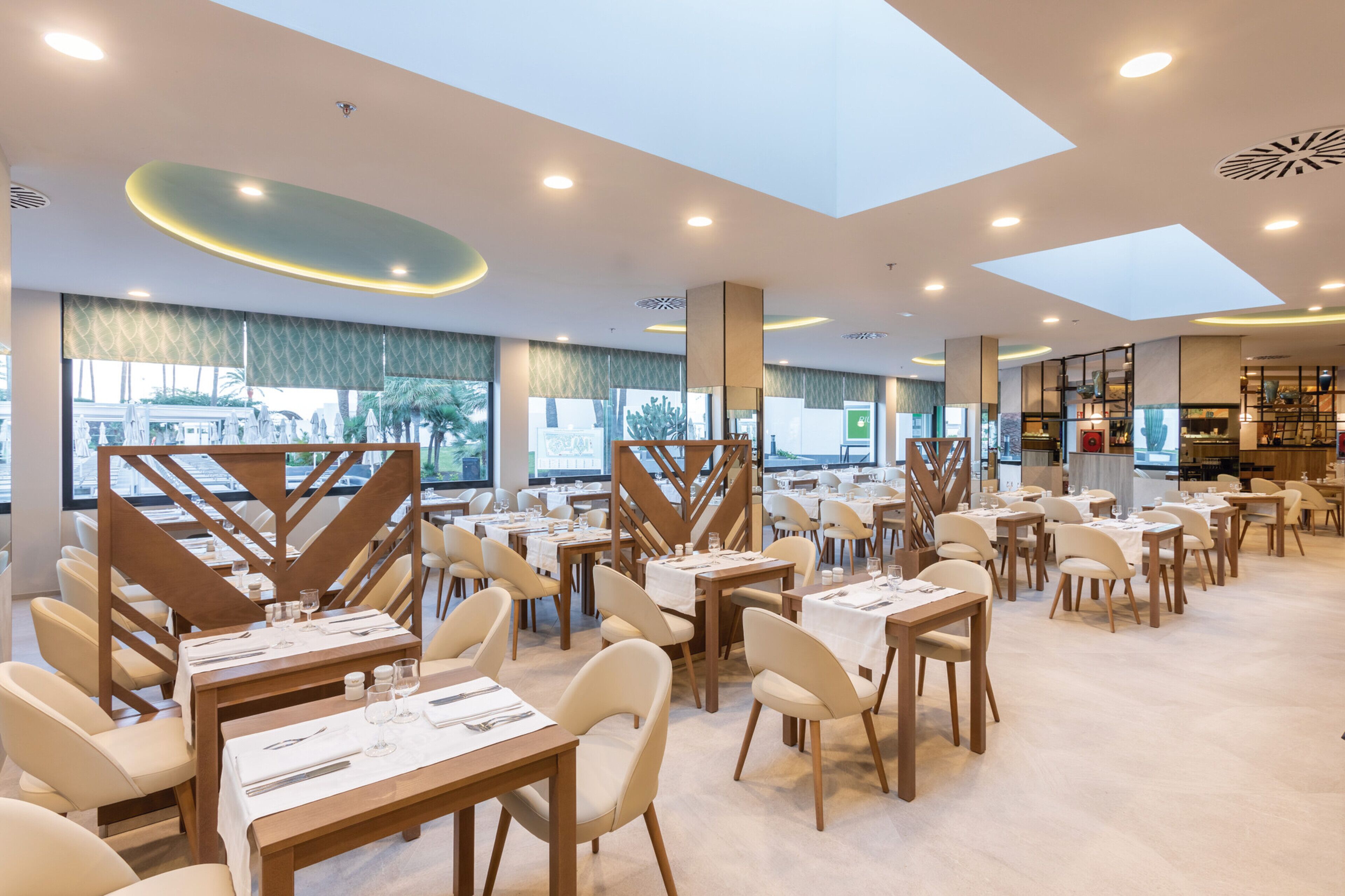 Un restaurante amplio con varias mesas y sillas, ventanas grandes y un diseño de techo moderno.