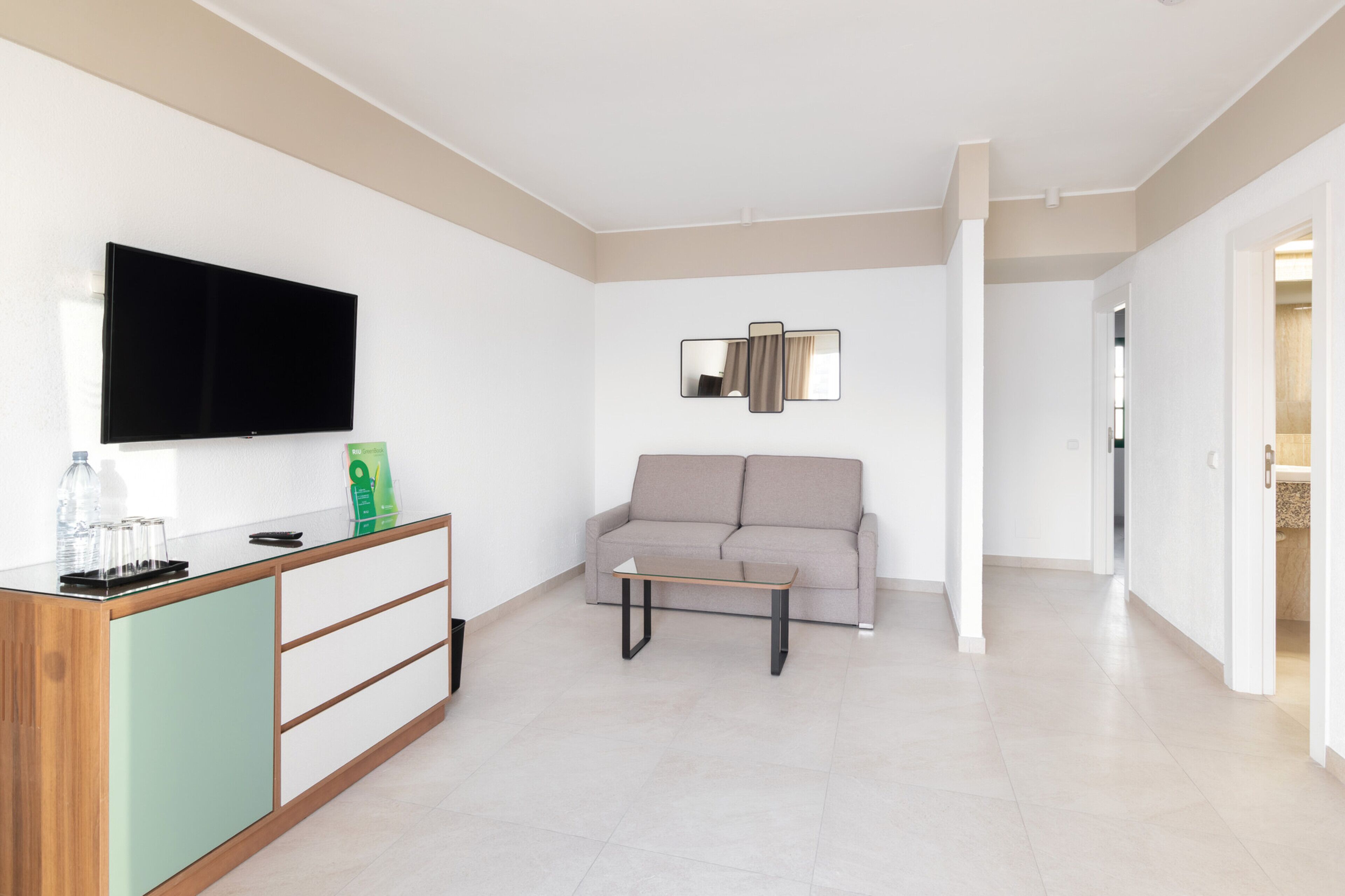 A modern living room with a flat-screen TV, a sofa, a coffee table, and a glass-top side table with a remote control and water bottles.