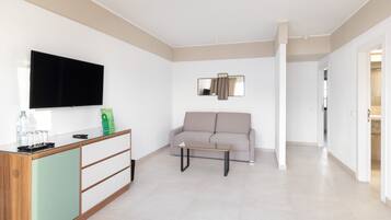A modern living room with a flat-screen TV, a sofa, a coffee table, and a glass-top side table with a remote control and water bottles.