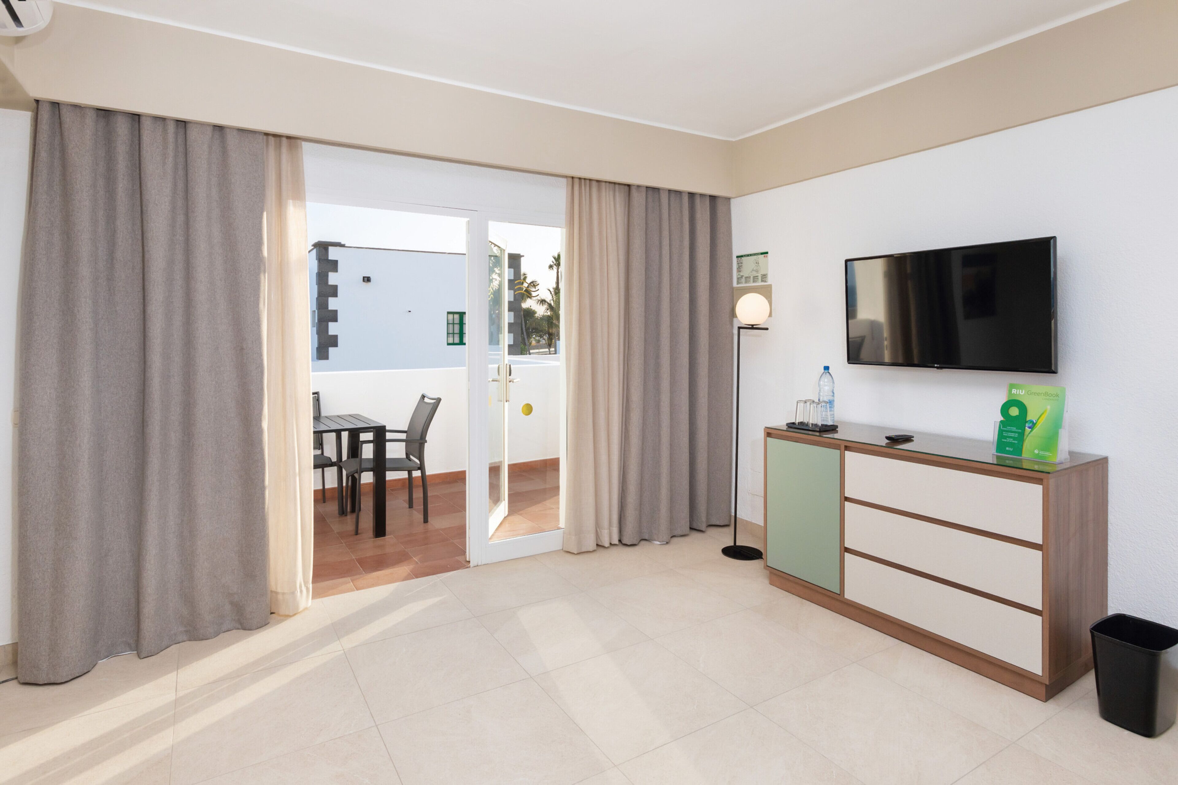 A hotel room with a TV, a small table, chairs, a glass cabinet, and a view through the door.