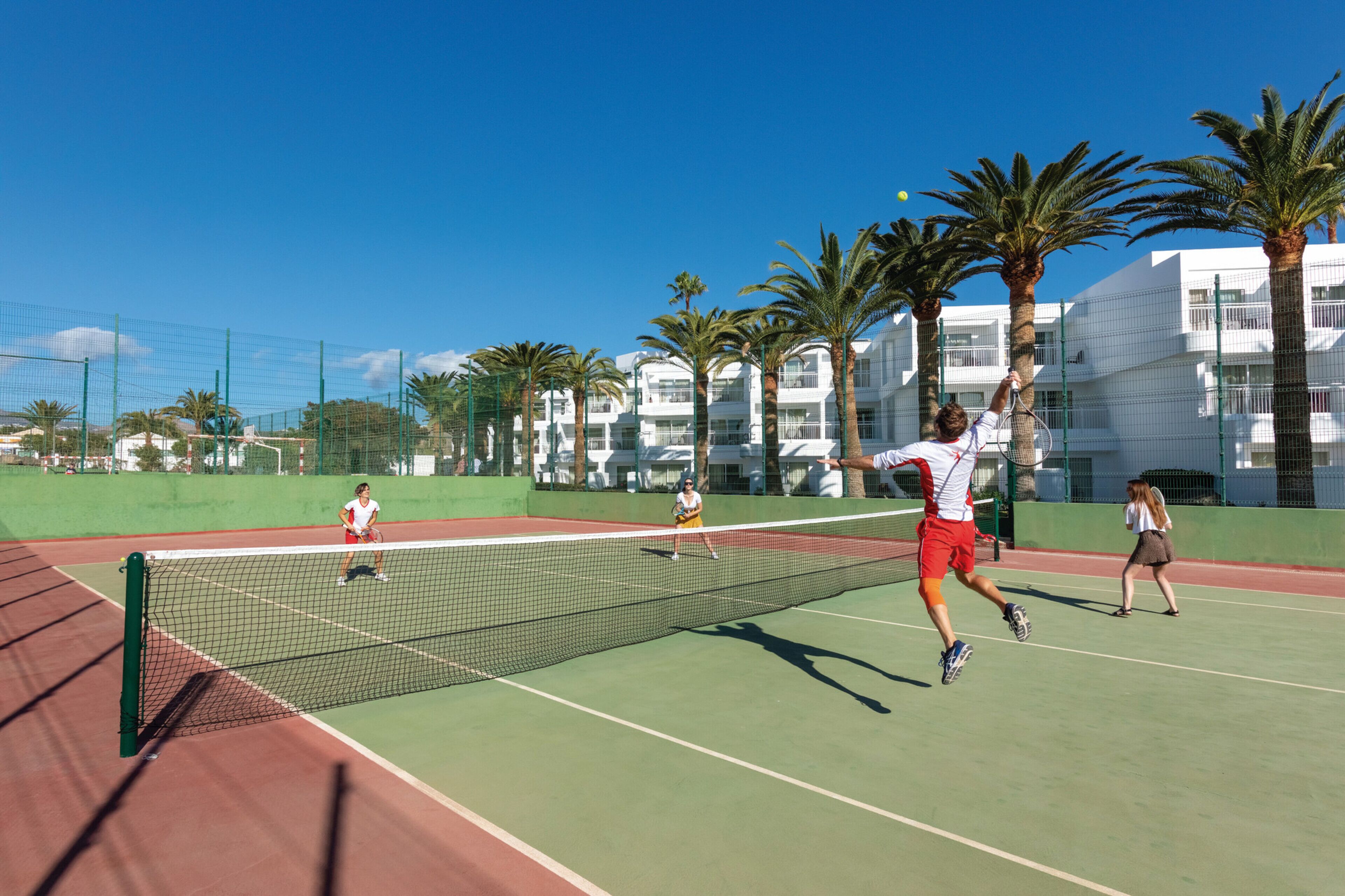 A tennis court with a net, palm trees, and a building in the background.