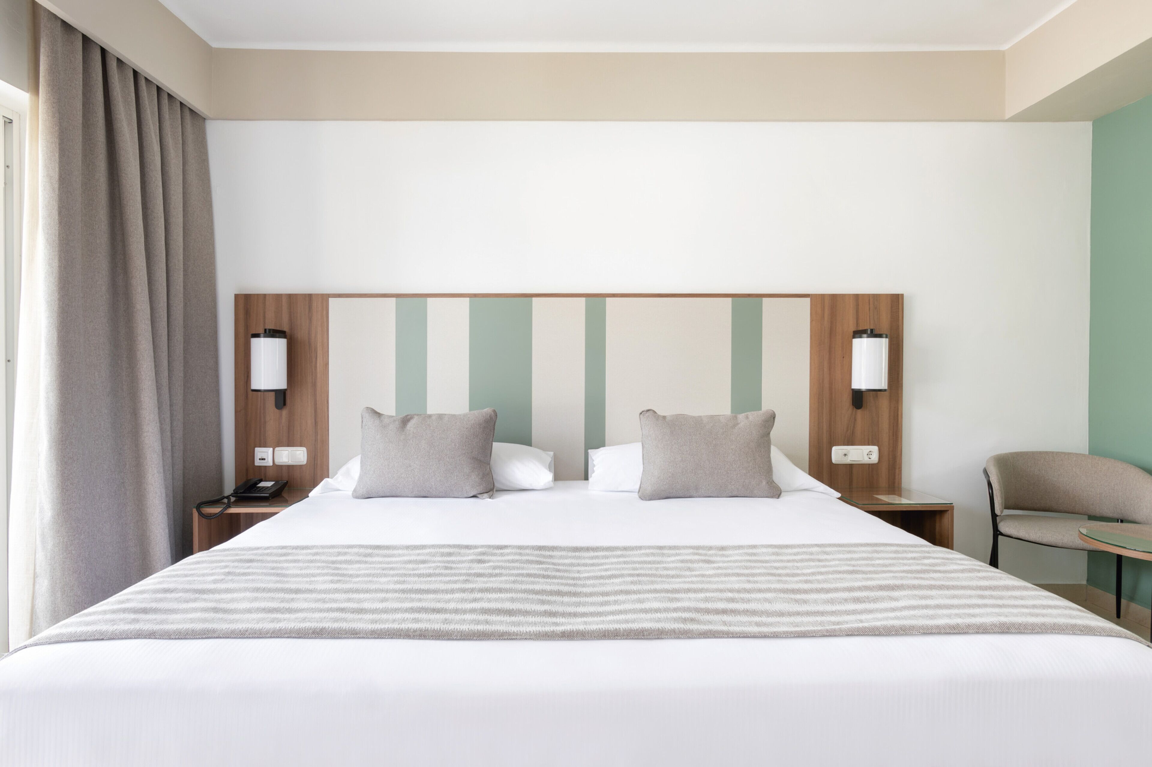 A hotel room with a large bed, two bedside tables, a chair, and a headboard with vertical stripes.
