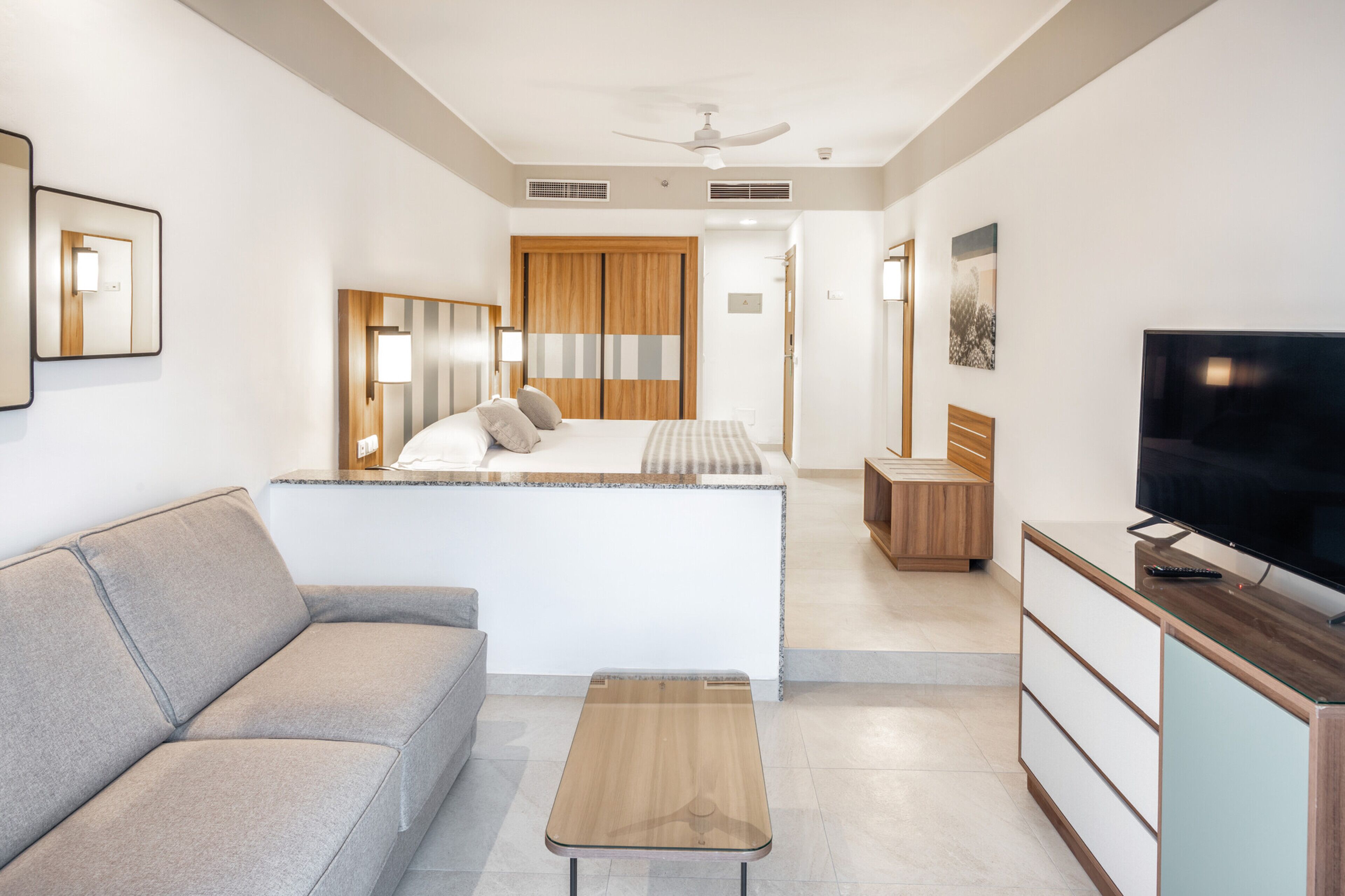 Una habitación de hotel moderna con sofá, una mesita, televisor y un armario de madera.