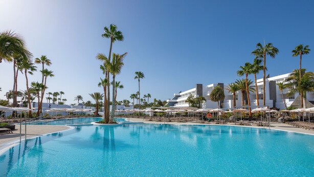 A large swimming pool with palm trees and sun loungers, surrounded by hotel buildings. - Hotel Riu Paraiso Lanzarote - All Inclusive (Tías)